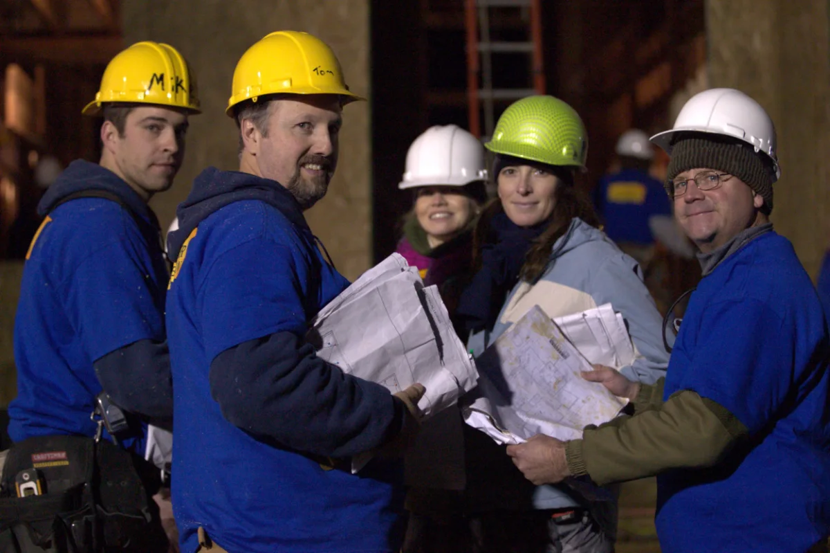 A framing crew and supervisors in hard hats on a job at dusk, holding a paper plan set