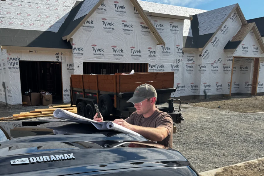 A superintendent at the hood of his pickup on a job site, plans spread out and his phone in hand, a Tyvek-wrapped house behind him