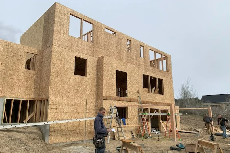 A small crew framing a three-storey house: one man in front with a tool belt, two more working beside the build