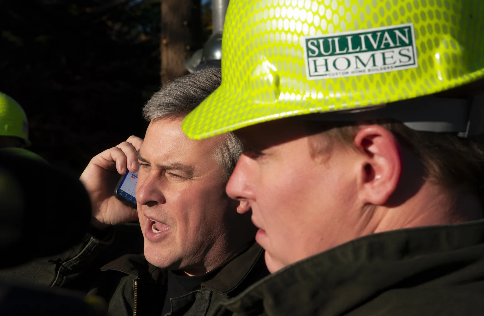 Todd Sullivan on a job site, phone to his ear, behind a crewman in a Sullivan Homes hard hat